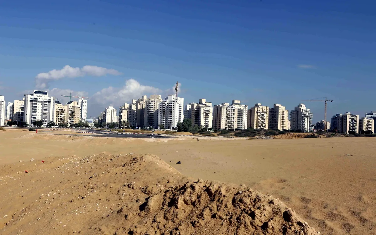 צילום של שטח חולי וערמת עפר מול שורת בנייני מגורים ועגורנים